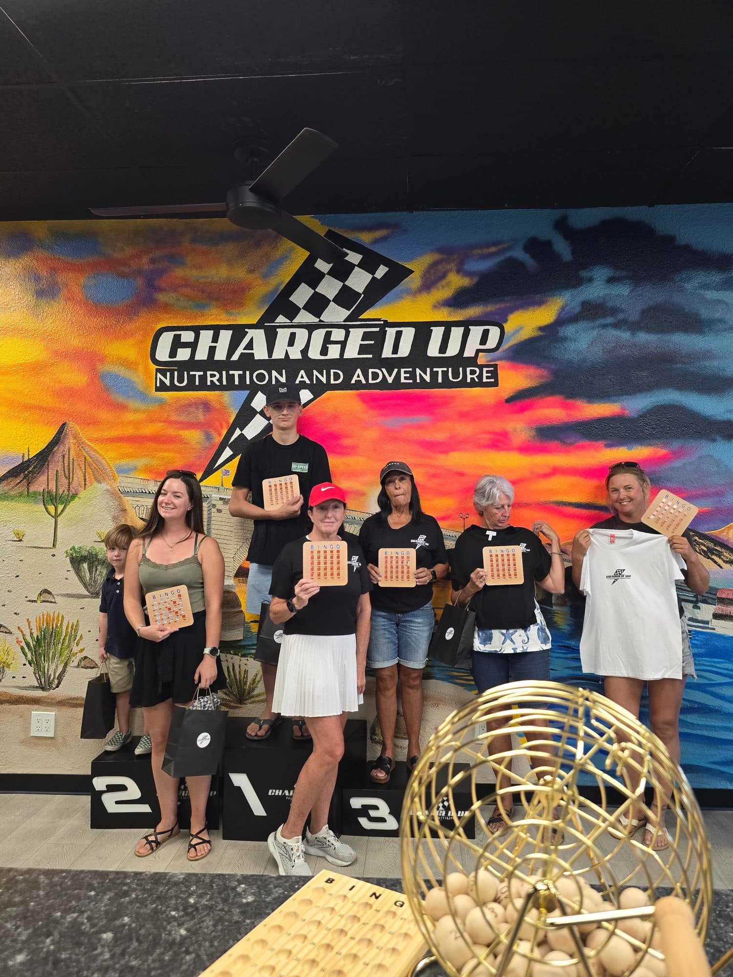 The Charged Up crew lined up on a numbered podium in front of the Charged Up Nutrition and Adventure mural, holding BINGO cards, with the bingo cage in front